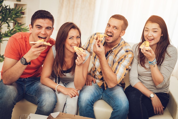 Quatre amis joyeux qui traînent dans un appartement. Ils mangent de la pizza et regardent la caméra.