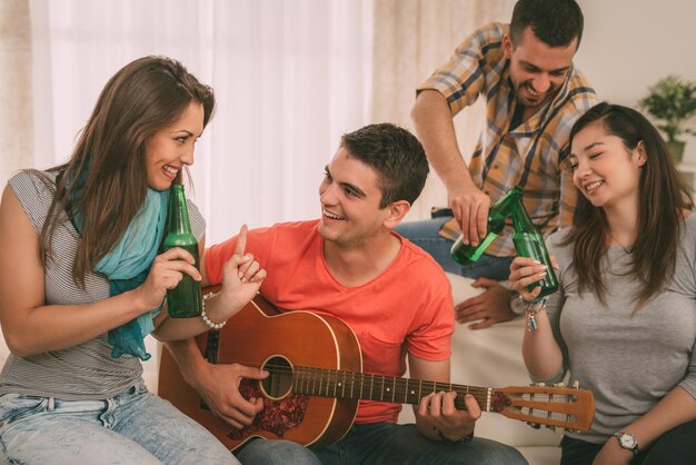 Quatre amis joyeux s'amusant avec de la guitare et de la bière dans un appartement.