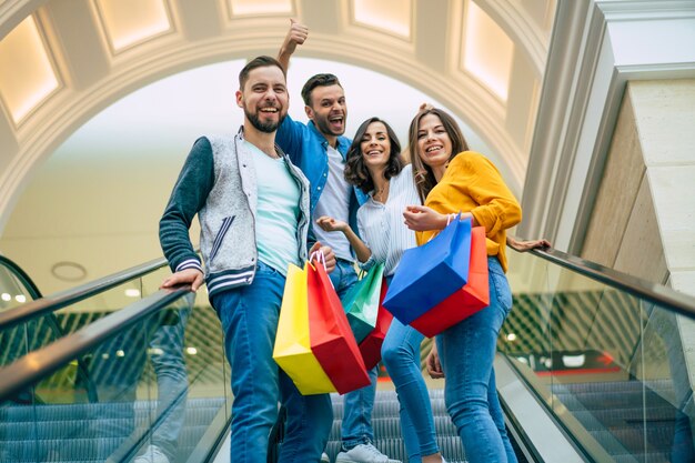 Quatre beaux amis modernes excités et souriant élégants dans des vêtements décontractés avec des sacs en papier s'amusent debout sur l'escalator dans le centre commercial pendant les achats.