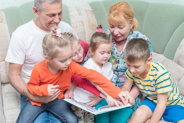 quatre enfants étudient un livre avec des grands-parents à la maison sur le canapé