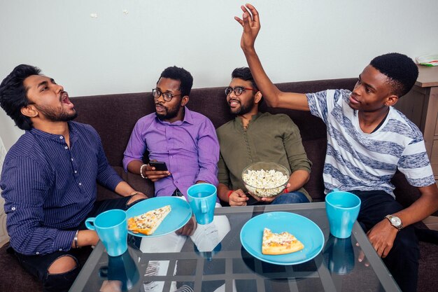 Quatre hommes afro-américains mangeant de la pizza à la maison en se jetant du pop-corn l'un dans l'autre