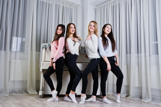 Quatre jolies filles amies portent des pulls chauds et des pantalons noirs contre un vieux piano avec une décoration de noël dans une salle blanche.