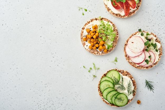 Quatre pains de sarrasin croustillants avec fromage à la crème, radis, tomate, pois chiche, concombre et microgreen pour un petit-déjeuner sain sur papier parchemin sur fond gris pierre. Concept végétalien et alimentation saine.