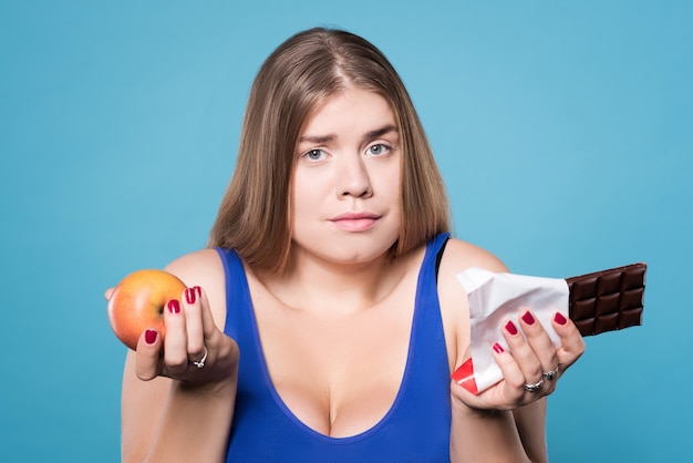 Que Choisir. Gros Plan D'une Belle Fille Dodue Confuse Tenant La Pomme Et Le Chocolat.