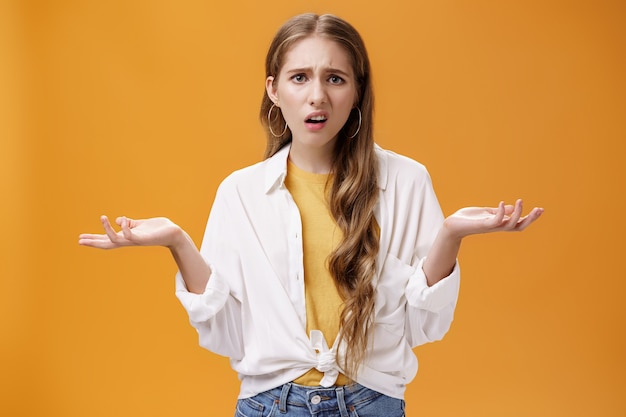 Ce que je ne comprends pas. Portrait d'une fille impolie interrogée arrogante, énervée par des questions haussant les mains, les mains écartées sur le côté, faisant une grimace irritée debout confuse sur le mur orange.