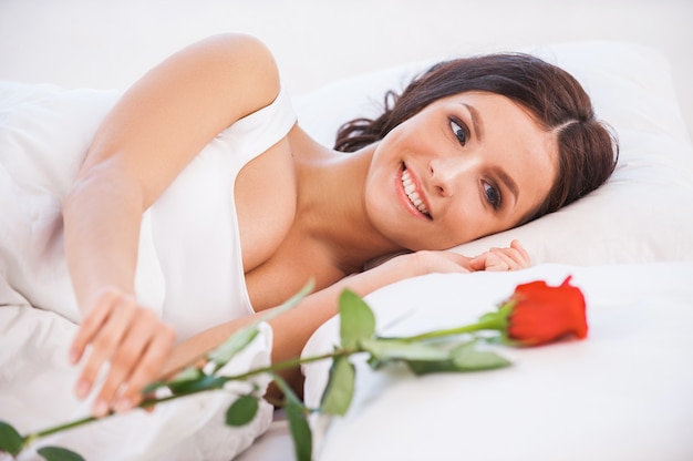 Quel beau matin. Vue latérale de la belle jeune femme au lit avec une rose rouge