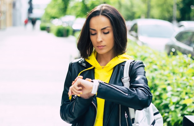 Quelle heure est-il? jeune femme, vêtue d'une veste en cuir et d'un sweat à capuche jaune, vérifie l'heure avec sa nouvelle smartwatch moderne.