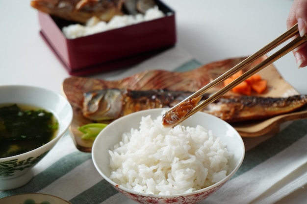 Quelqu'un à l'aide de baguettes essayant de choisir un morceau de poisson saba ou maquereau grillé servi avec du riz cuit et de la soupe miso sur un napperon rayé blanc et vert sur un tableau blanc