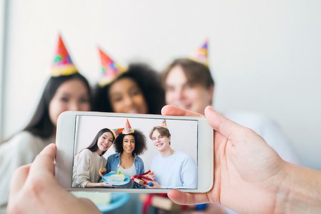 Quelqu'un prend une photo au téléphone. Il y a une fille qui fête son anniversaire et ses amis qui sont réunis. Ils posent et sourient à la caméra.