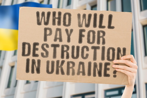 La question Qui paiera pour la destruction en Ukraine est sur une bannière entre les mains des hommes avec un drapeau ukrainien flou en arrière-plan Ville Russie Rue Sponsor Attaque Guerre Danger Urbain