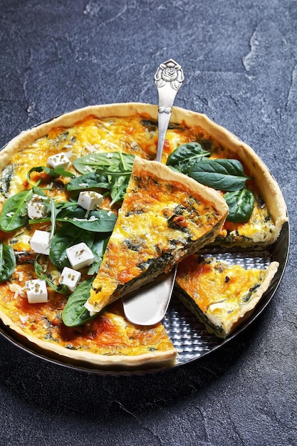 Quiche au fromage cheddar et épinards feta dans un plat allant au four sur une planche de bois sur une table en béton gris avec une tranche sur une spatule, vue verticale d'en haut, cuisine française