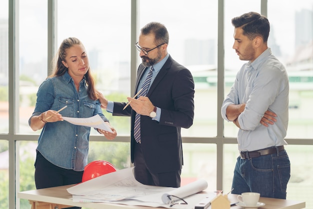 Équipe d'architectes multiethniques travaillant sur les plans de construction dans la salle de réunion. Ingénieurs discutant d'un projet au bureau. Homme d'affaires mature et femme debout autour de la table travaillant sur le plan.