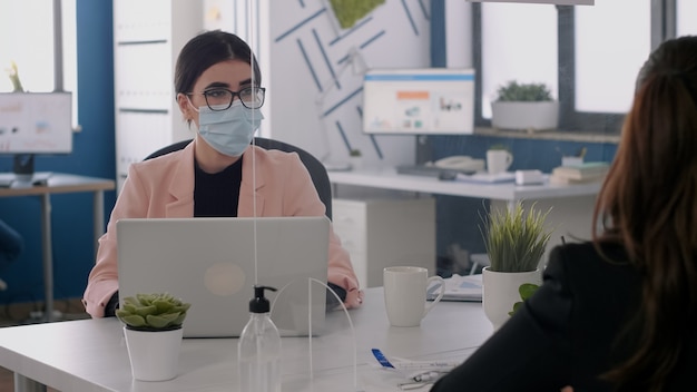 Équipe commerciale avec des masques faciaux travaillant sur des idées de marketing tout en regardant un écran d'ordinateur assis dans un nouveau bureau normal. Collègues respectant la distanciation sociale pendant la pandémie mondiale de coronavirus