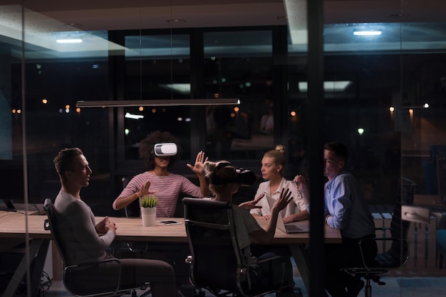 Équipe commerciale multiethnique utilisant un casque de réalité virtuelle lors d'une réunion de bureau de nuit Les développeurs se réunissent avec un simulateur de réalité virtuelle autour d'une table dans un bureau créatif.