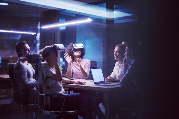 Équipe commerciale multiethnique utilisant un casque de réalité virtuelle lors d'une réunion de bureau de nuit Les développeurs se réunissent avec un simulateur de réalité virtuelle autour d'une table dans un bureau créatif.