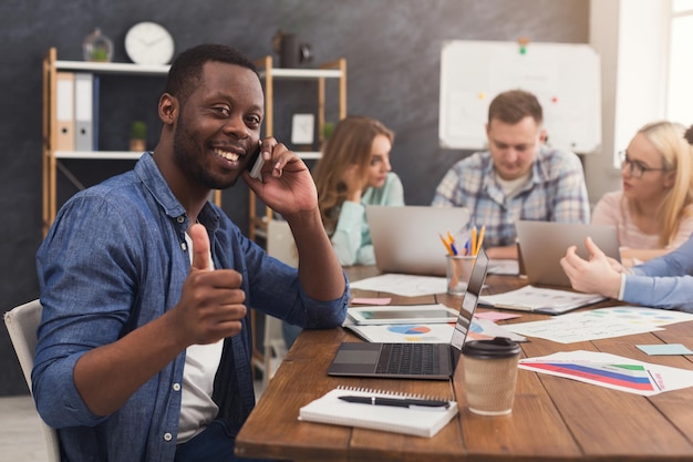 Équipe commerciale parlant des résultats de leur travail. Groupe d'hommes d'affaires joyeux discutant d'une nouvelle stratégie marketing, jeune homme noir parlant au téléphone et montrant le pouce vers le haut, espace de copie