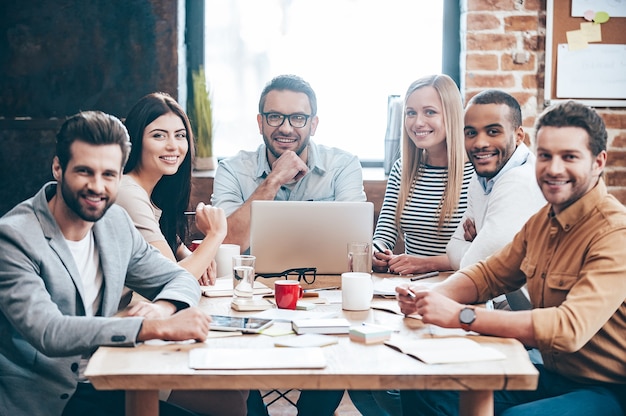 Équipe créative. Groupe de six jeunes joyeux regardant la caméra avec le sourire alors qu'ils étaient assis à la table au bureau