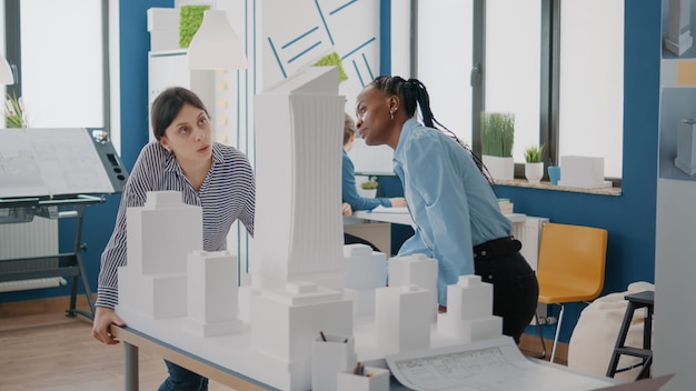 Équipe de femmes multiethniques analysant une maquette sur une table pour concevoir un modèle de construction. Les collègues réfléchissent ensemble à des idées pour planifier la structure de la construction urbaine et la disposition architecturale.