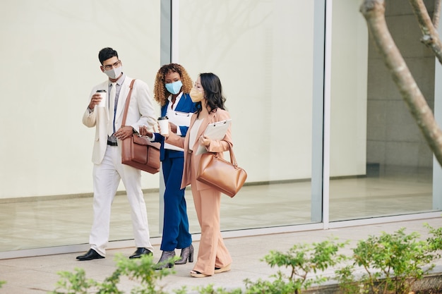 Équipe d'hommes d'affaires confiants et prospères marchant dans la rue avec des masques de protection, buvant du café et discutant du travail