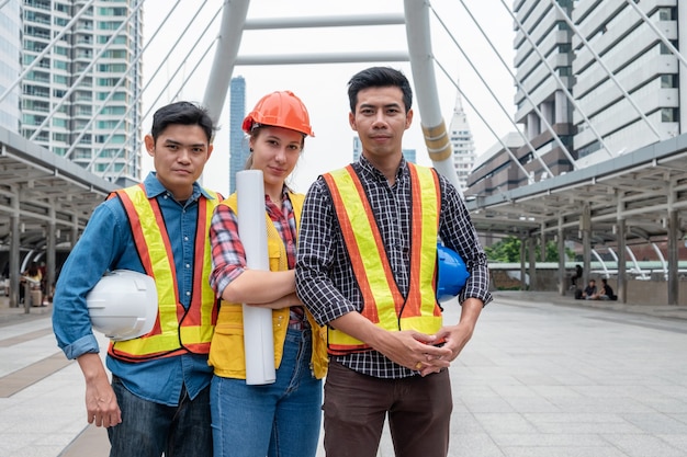 Équipe d'ingénieurs multiethniques confiant debout et tenant un plan, casque de sécurité sur un chantier de construction en milieu urbain