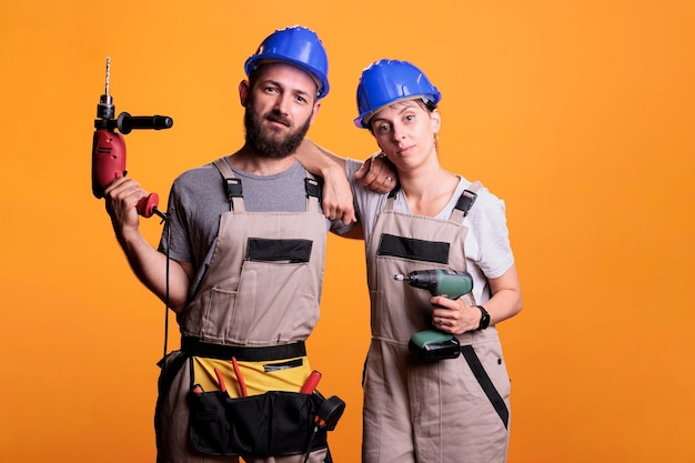 Équipe d'ingénieurs professionnels tenant des perceuses électriques en studio, posant avec des outils électriques sur fond. Homme et femme utilisant un pistolet de forage et portant une combinaison avec un casque, une ceinture à outils.