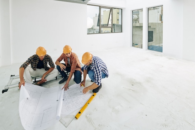 Équipe de jeunes constructeurs dans des casques de vérification des plans avant de commencer le processus de rénovation dans l'appartement du client