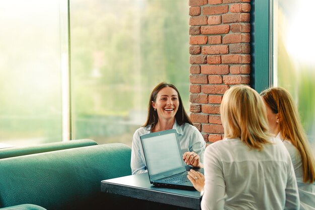 Équipe réussie. Trois femme d'affaires confiante porte des chemises blanches se regardant et souriant alors qu'il était assis au bureau au bureau avec un ordinateur portable sur le bureau.