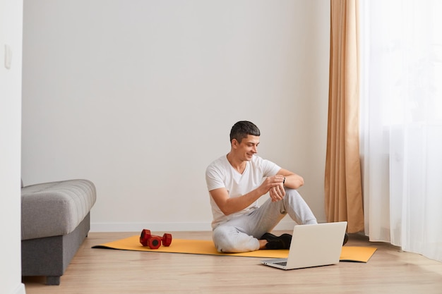 Équipement sportif et activités à la maison Guy avec tracker de fitness assis sur le sol sur un tapis en regardant les haltères de l'écran de l'appareil près de lui à l'intérieur du salon en suivant son activité