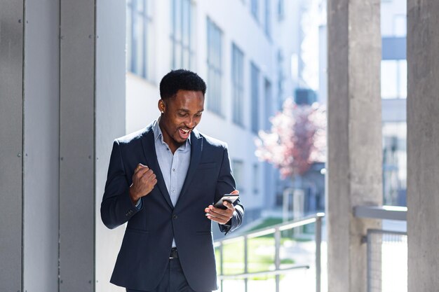 A quitté l'homme d'affaires afro-américain célébrant ses réalisations en regardant un smartphone. Victoire réussie d'un homme d'affaires avec un téléphone portable à l'extérieur. Happy Good News Success Deal, taux de réussite de Startup Bet