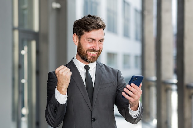 A quitté l'homme d'affaires célébrant les réalisations en regardant l'extérieur du smartphone. Homme d'affaires prospère célébrant la victoire avec un téléphone portable à l'extérieur. Bonne Bonne Nouvelle. Accord réussi, pari de démarrage, commerce