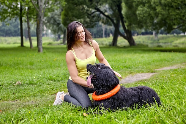 Race de chien noir Briard allongé sur l'herbe et regarde sa propriétaire, qui est assise à proximité.