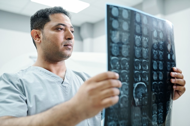 Radiologue métis contemporain en uniforme regardant l'image radiographique de l'un des patients et l'analysant par rapport à l'environnement du cabinet médical