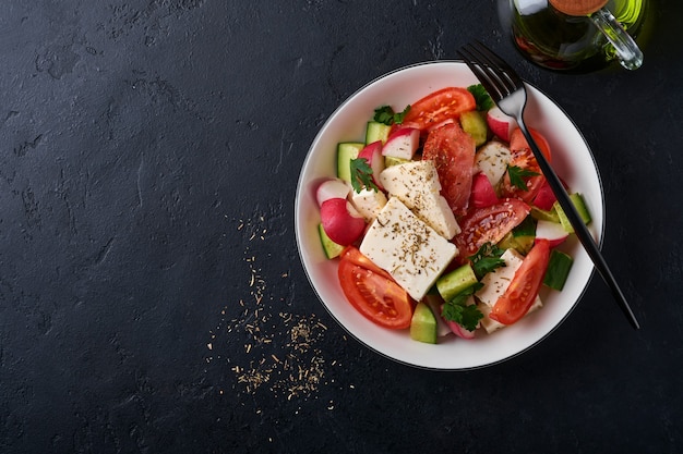 Radis, concombre, tomate, poivre et fromage feta aux épices, poivre et huile d'olive dans un bol blanc sur fond d'ardoise noire, de pierre ou de béton. Concept d'alimentation saine. Vue de dessus.