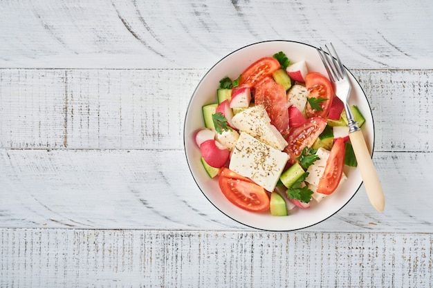 Radis, concombre, tomate, poivre et fromage feta avec épices poivre et huile d'olive dans un bol blanc sur fond gris ardoise, pierre ou béton. Concept d'alimentation saine. Vue de dessus.