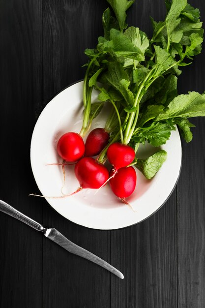 Radis frais dans une assiette blanche et un couteau sur un fond en bois foncé. Vue de dessus. Contexte alimentaire.