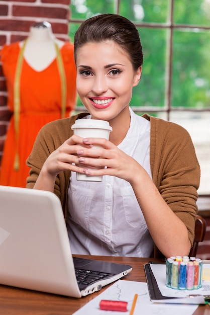 Rafraîchir son esprit. Jeune créatrice de mode joyeuse tenant une tasse avec une boisson chaude et souriante assise sur son lieu de travail et avec un mannequin debout en arrière-plan