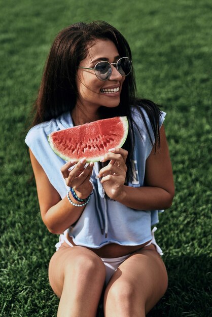 Rafraîchissement d'été. Jolie jeune femme en lunettes tenant une tranche de pastèque et souriante assise sur l'herbe