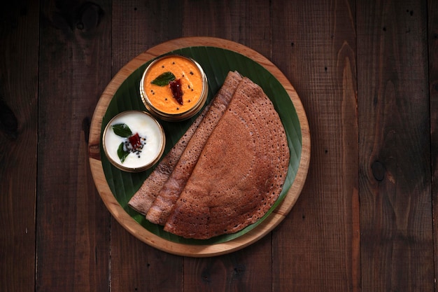 Ragi Dosa, petit-déjeuner sain de l'Inde du Sud disposé sur une base ronde en bois bordée de feuilles de bananier et de chutney de noix de coco placé à côté.