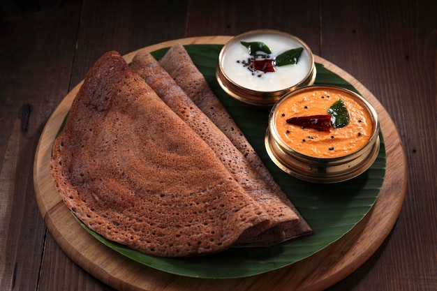 Ragi Dosa, petit-déjeuner sain de l'Inde du Sud disposé sur une base ronde en bois bordée de feuilles de bananier et de chutney de noix de coco placé à côté.