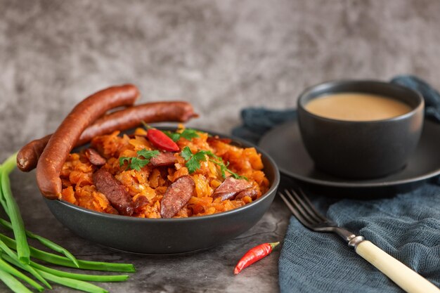 Ragoût de chou dans un bol avec des saucisses fumées et une tasse de café sur fond gris. Vue de côté.