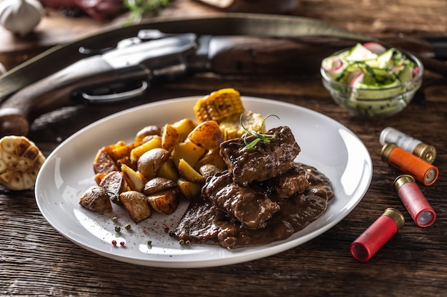 Ragoût de goulasch de gibier sauvage servi avec des pommes de terre rôties et assaisonné de grains de poivre de quatre couleurs. La nourriture entière est entourée de fusils de chasse et de balles de fusil.