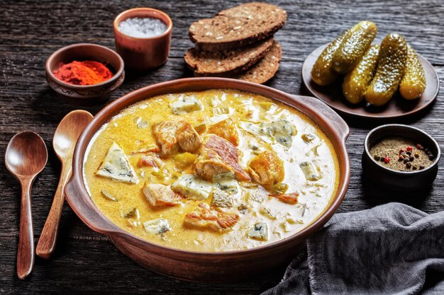 Photo ragoût de porc avec crème, cornichons et fromage à moisissure bleue cuit de filet de porc maigre avec paprika fumé et poivre noir servi sur un pot en argile avec une cuillère en bois sur une table en bois sombre, vue de dessus, gros plan