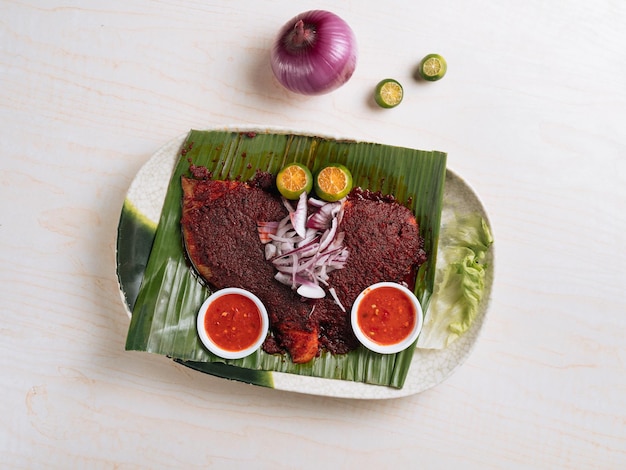 Raie barbecue traditionnelle servie sur une feuille de bananier avec des oignons, de la sauce aux piments rouges et du citron sur une assiette blanche vue de dessus