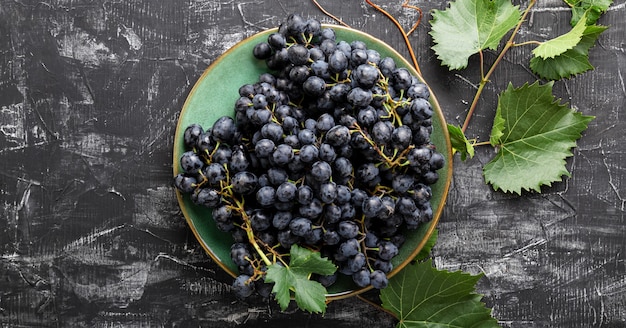 Raisins noirs en plaque avec plante de vigne sur la vue de dessus de table sombre. Nourriture sucrée de raisins juteux sur fond de béton noir à plat. Bannière Web longue.