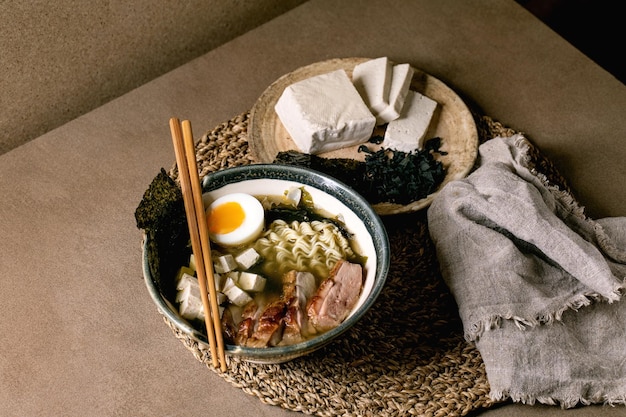 Photo ramen de soupe de style asiatique fait maison avec nouilles grillées magret de canard tofu chips de nori aux algues et œuf à la coque dans un bol en céramique avec des baguettes et une cuillère sur une table de cuisine marron