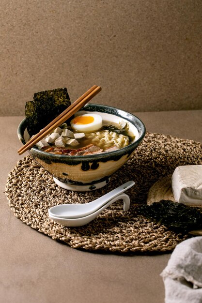 Ramen de soupe de style asiatique fait maison avec nouilles grillées magret de canard tofu chips de nori aux algues et œuf à la coque dans un bol en céramique avec des baguettes et une cuillère sur une table marron avec une serviette en paille tricotée