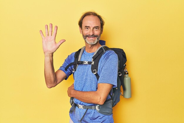 Randonneur d'âge moyen avec sac à dos en studio souriant joyeux montrant le numéro cinq avec les doigts