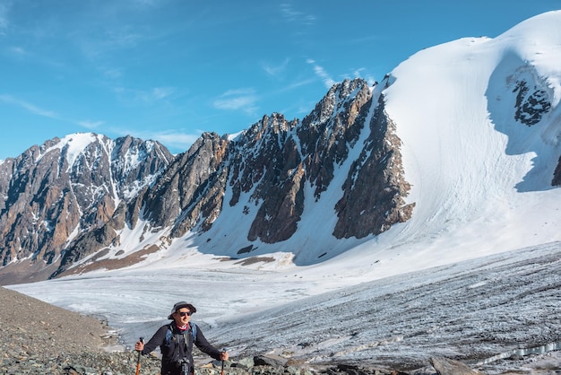 Un randonneur avec des bâtons de trekking se promène près d'une grande langue glaciaire dans les hautes montagnes Paysage pittoresque avec un touriste près d'un glacier au soleil Homme avec des lunettes de soleil et un appareil photo parmi les montagnes enneigées en journée ensoleillée