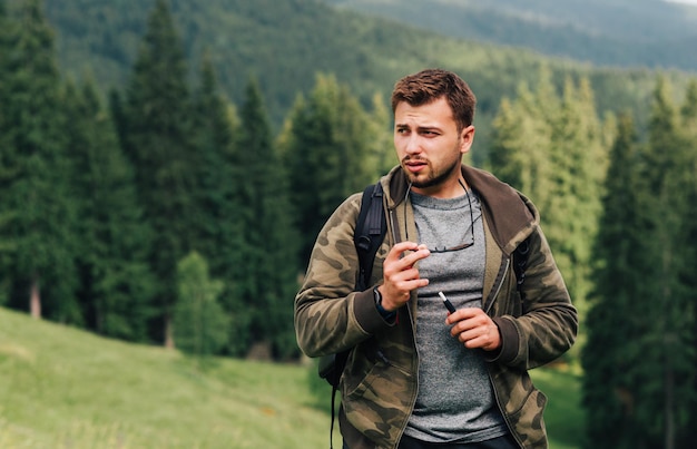 Randonneur mâle adulte avec une cigarette électronique dans ses mains se tient dans les montagnes et regarde sur le côté sur fond d'une vue irréelle sur les montagnes