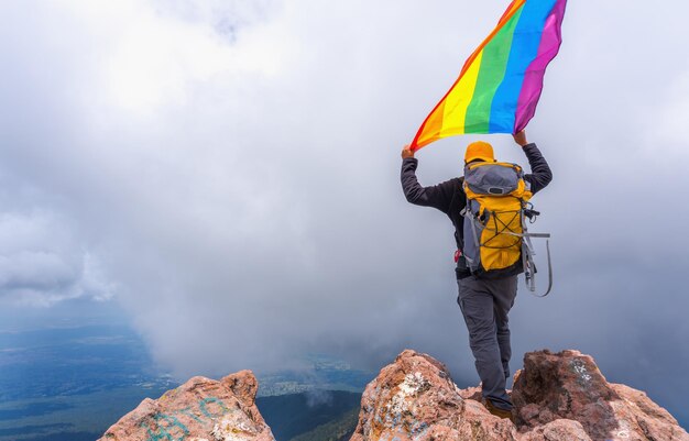 Un randonneur avec un sac à dos debout au sommet de la montagne et tenant un drapeau de fierté